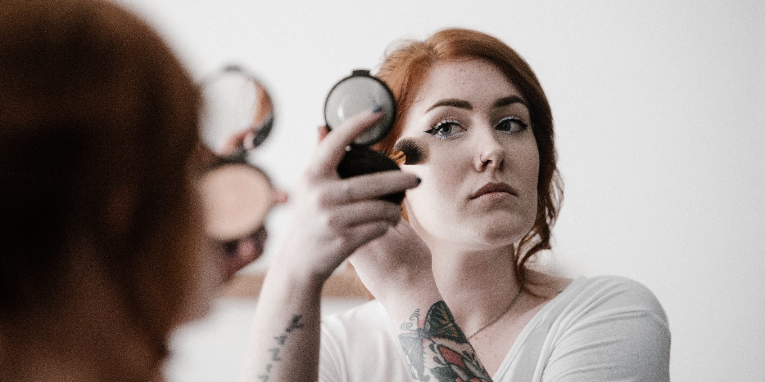 femeie, tatuaje, machiaj, oglinda O femeie cu tatuaje se machiaza cu o pensula in timp ce se uita in oglinda.
