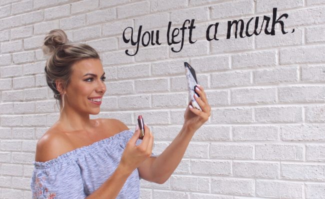 O fata care tine un ruj se admira in oglinda, pe fundalul un perete pe care scrie You left a mark.